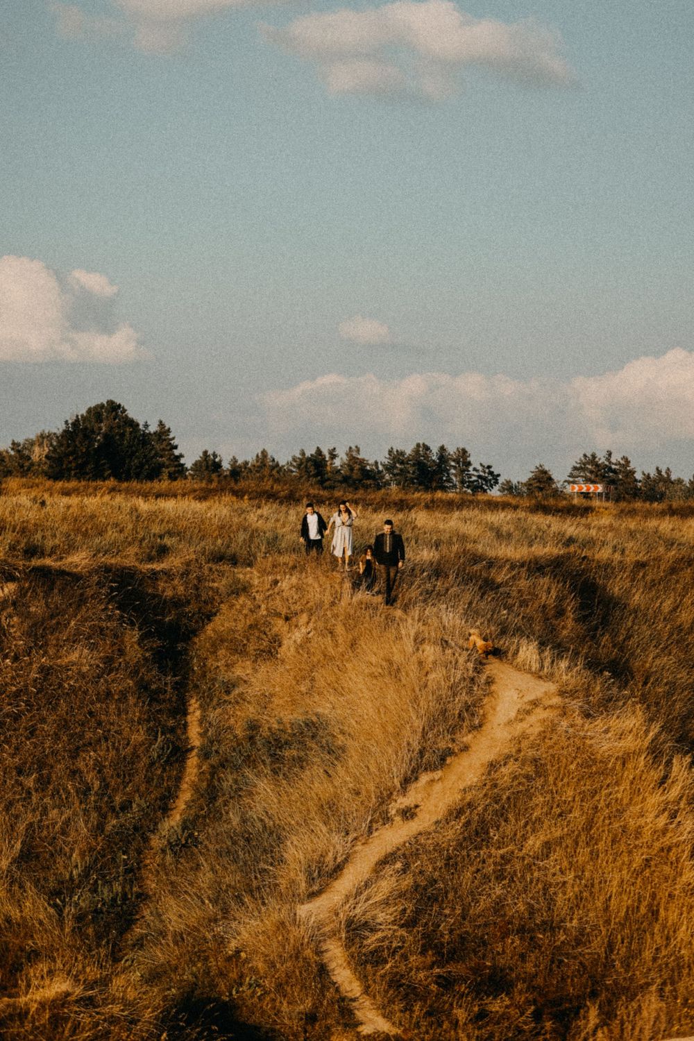 People walking along a winding dirt trail through golden grassy hills under a pale blue sky with scattered clouds.