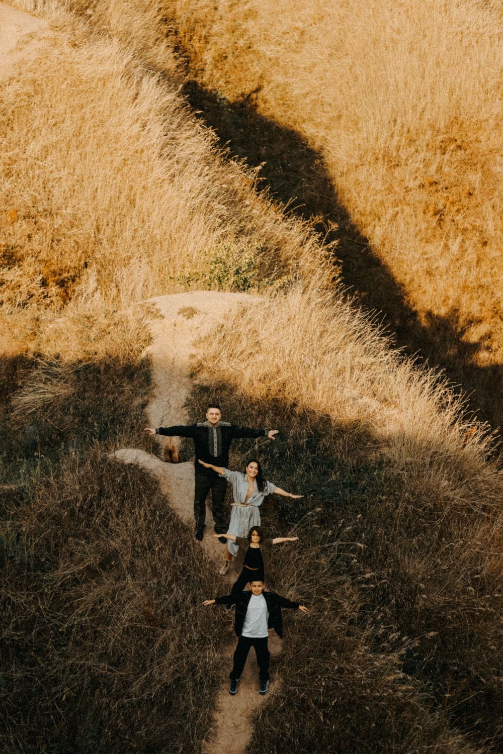 Three people stand with outstretched arms on a hiking trail surrounded by golden dry grass on a hillside at sunset.