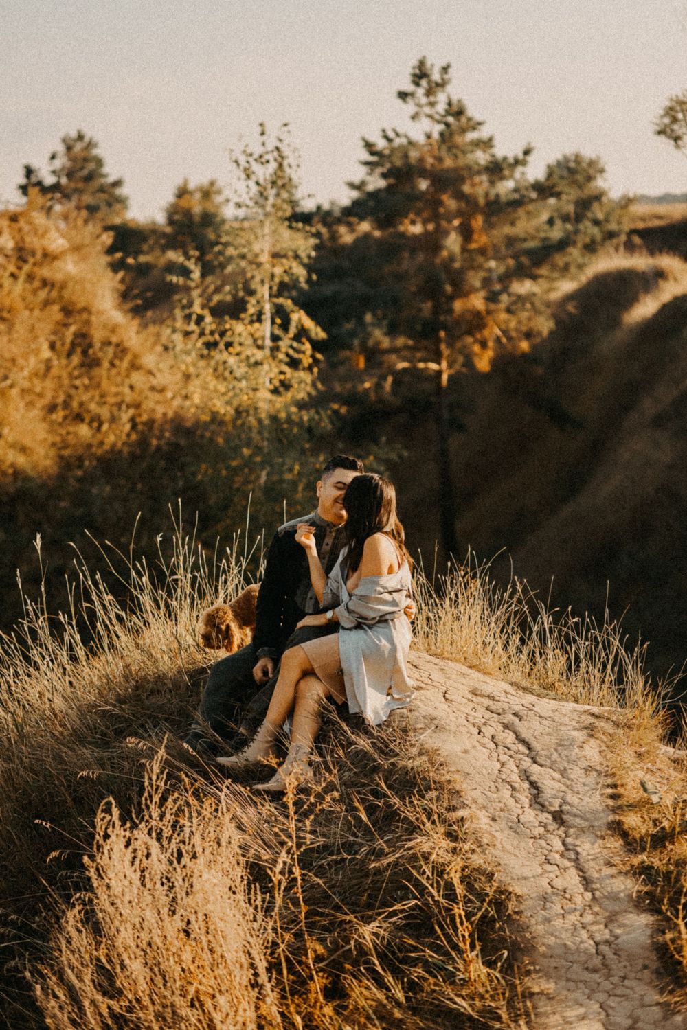 A couple sits together on a dirt path at golden hour with sunlit pine trees and tall grass in the background.