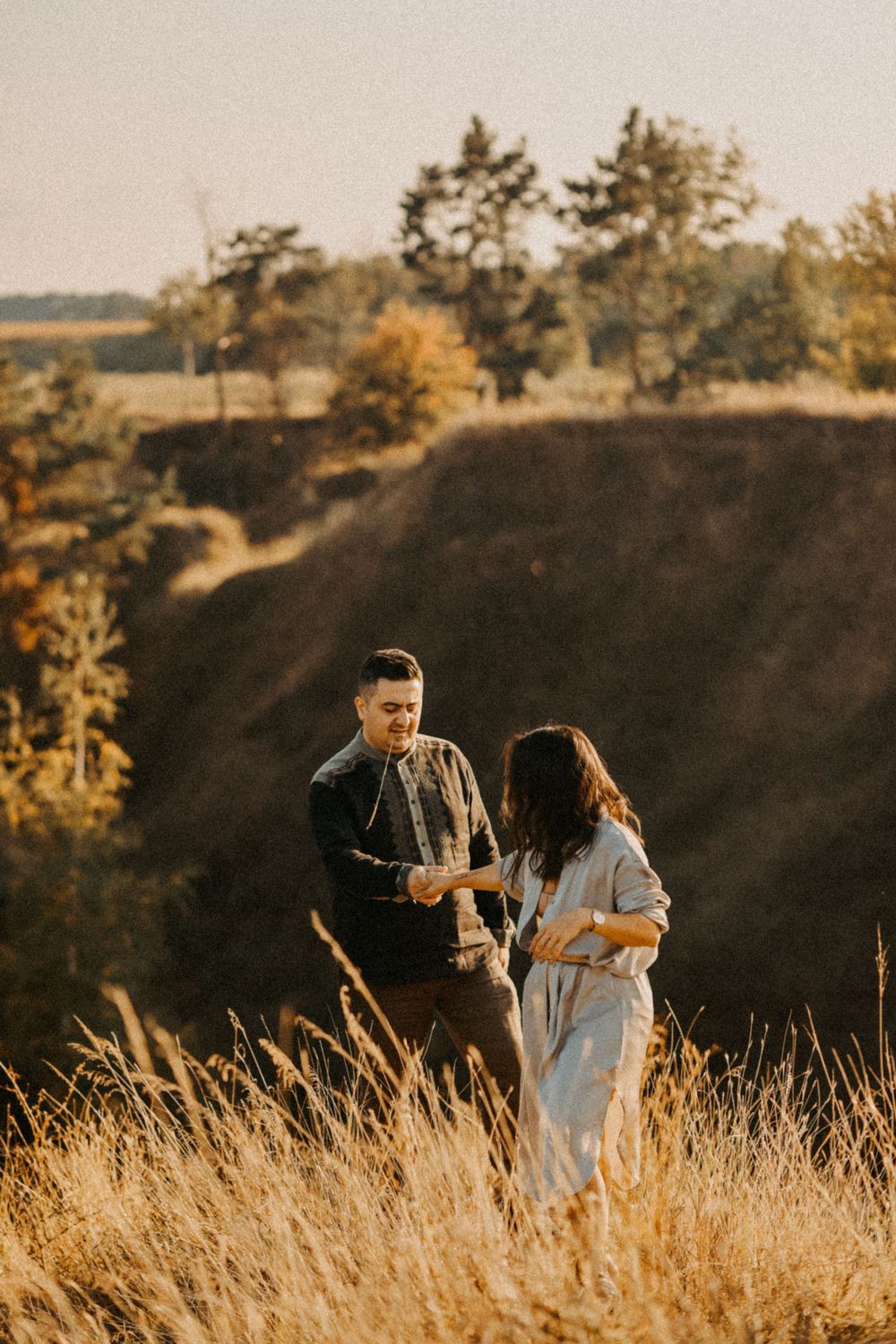 Two people stand in tall golden grass at sunset on a hillside silhouetted against warm evening light.