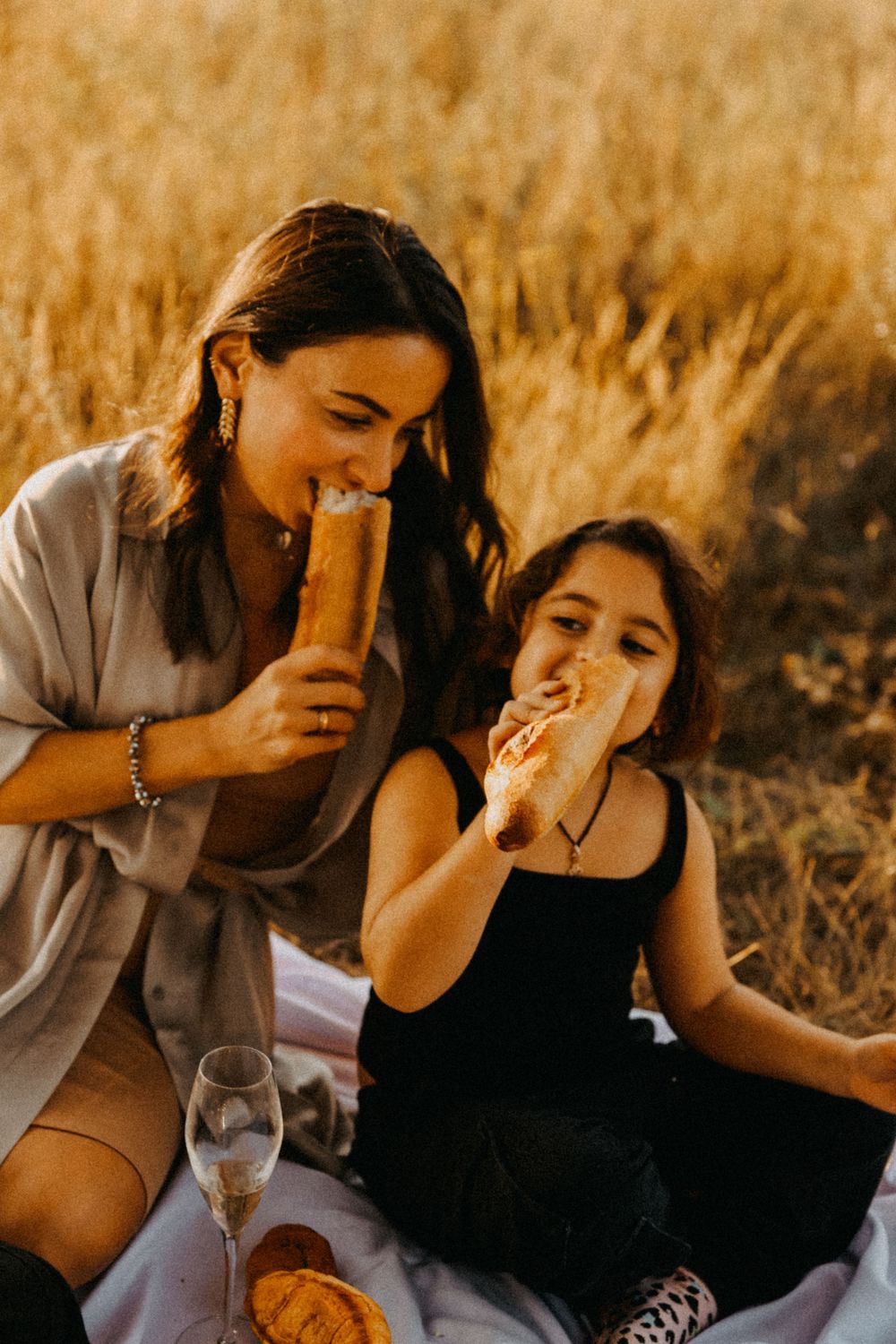 Two people enjoy fresh baguettes and wine during a golden hour picnic in a wheat field.