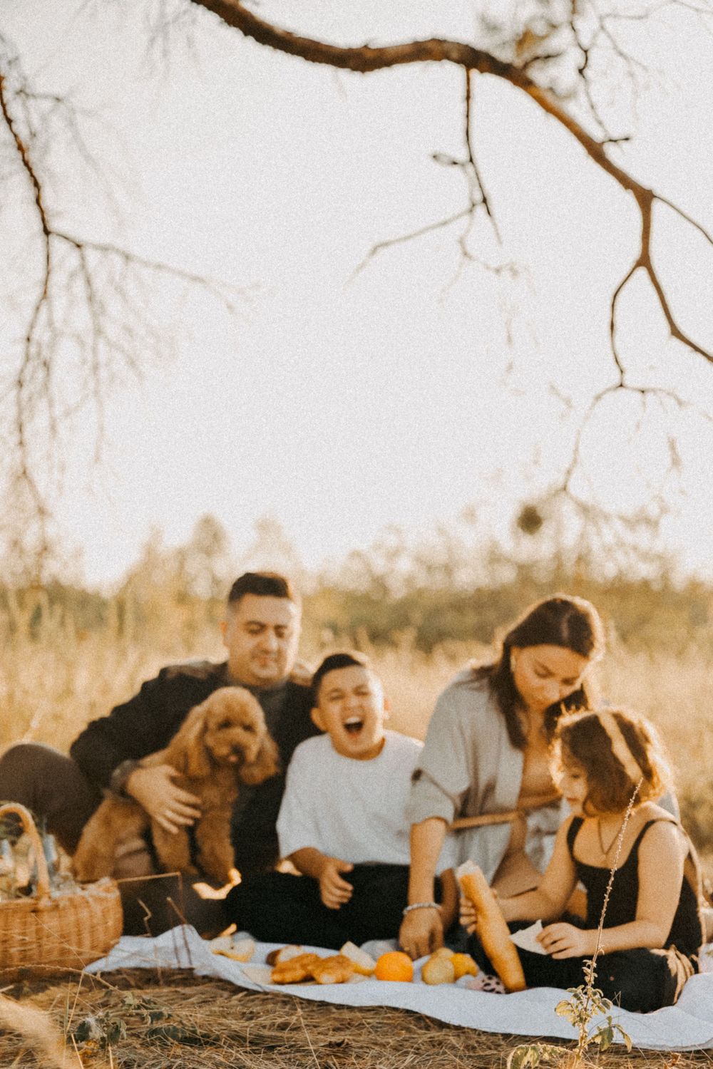 A group enjoying a casual outdoor picnic on a blanket with a golden retriever puppy and basket under a tree branch.