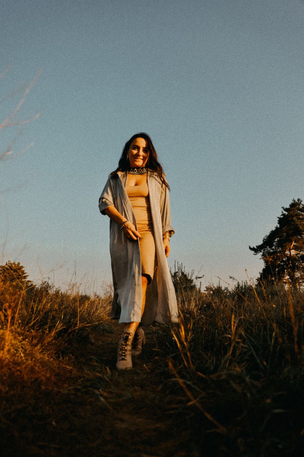 Someone in a long beige coat and dress stands in a field at golden hour with warm sunset lighting.