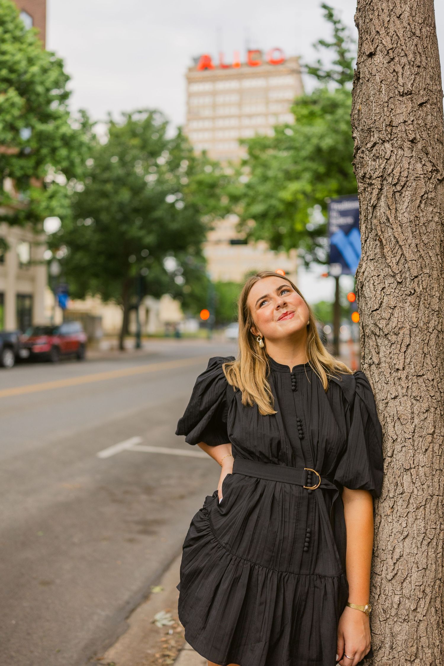 Young woman in black puffed-sleeve dress leaning against tree on urban street with Alico building in background.