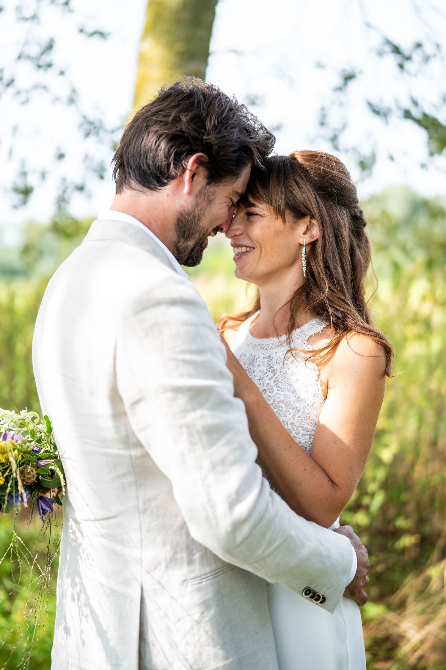 Romantic wedding couple in white attire embrace outdoors in natural garden setting with soft sunlight.