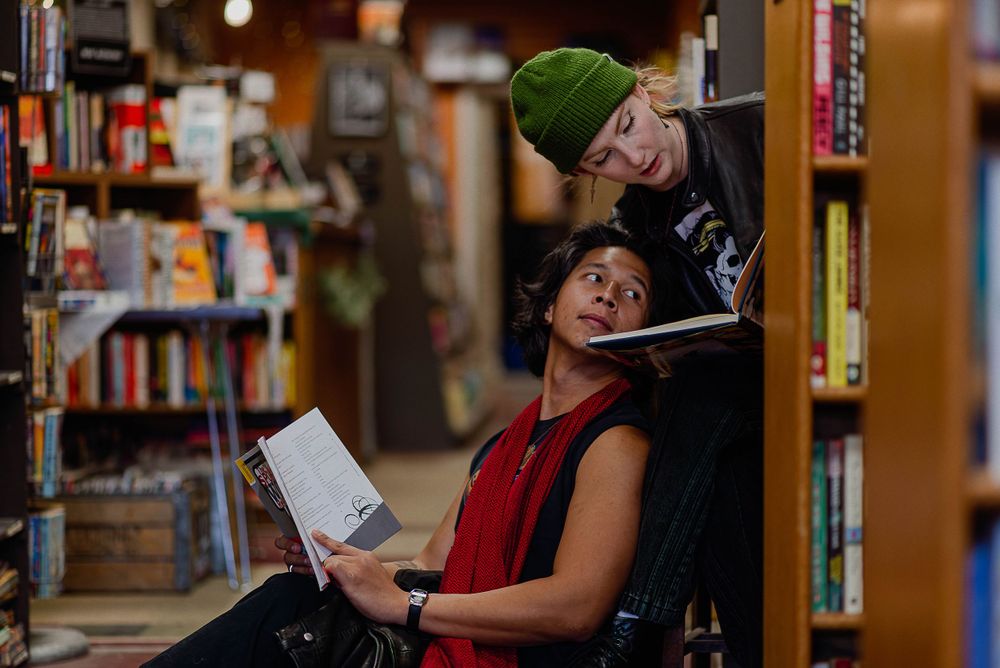 Two people browse books together in a cozy library aisle lined with bookshelves and warm lighting.