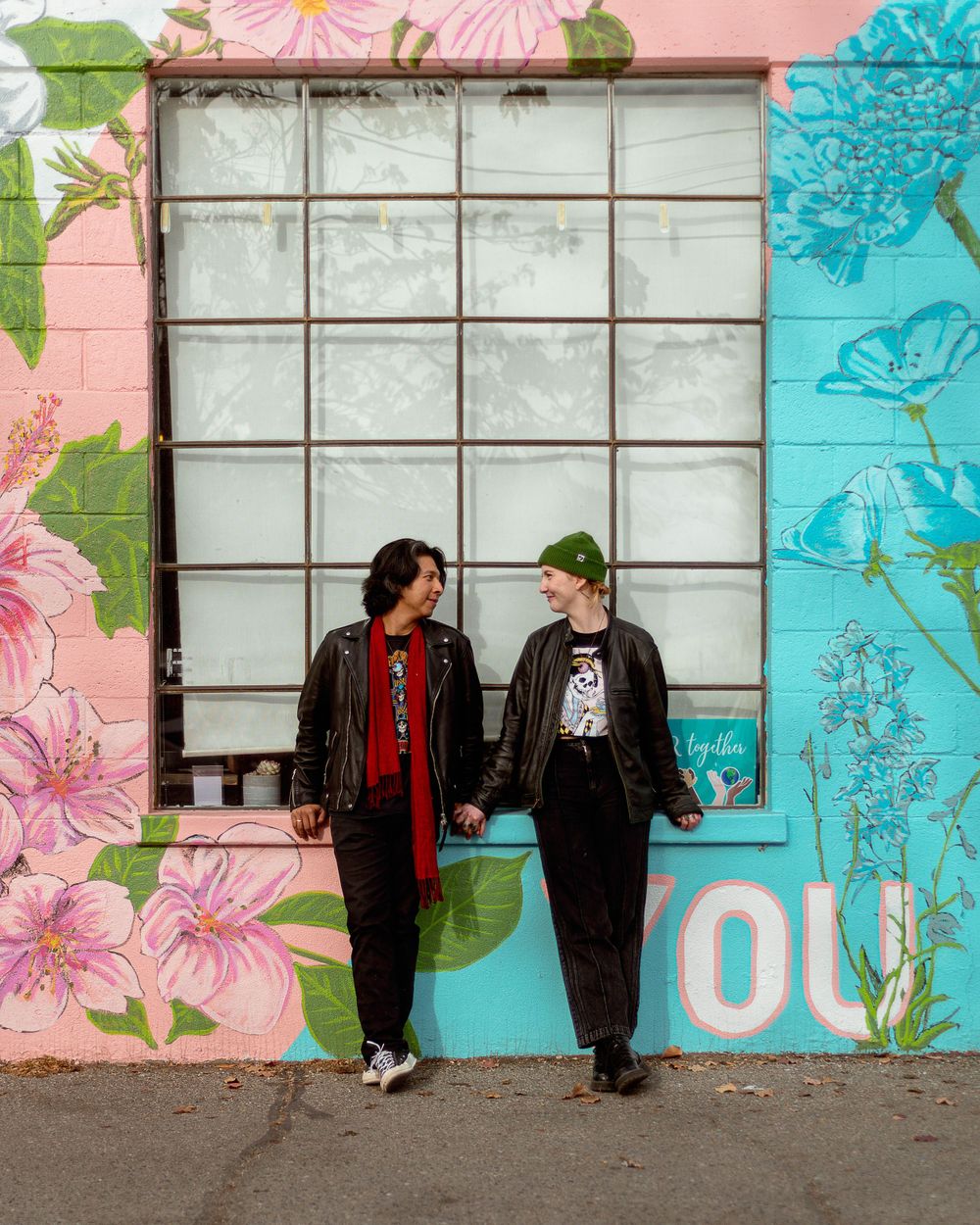 Two people lean against a wall with pink and turquoise floral murals, wearing black jackets in an urban setting.