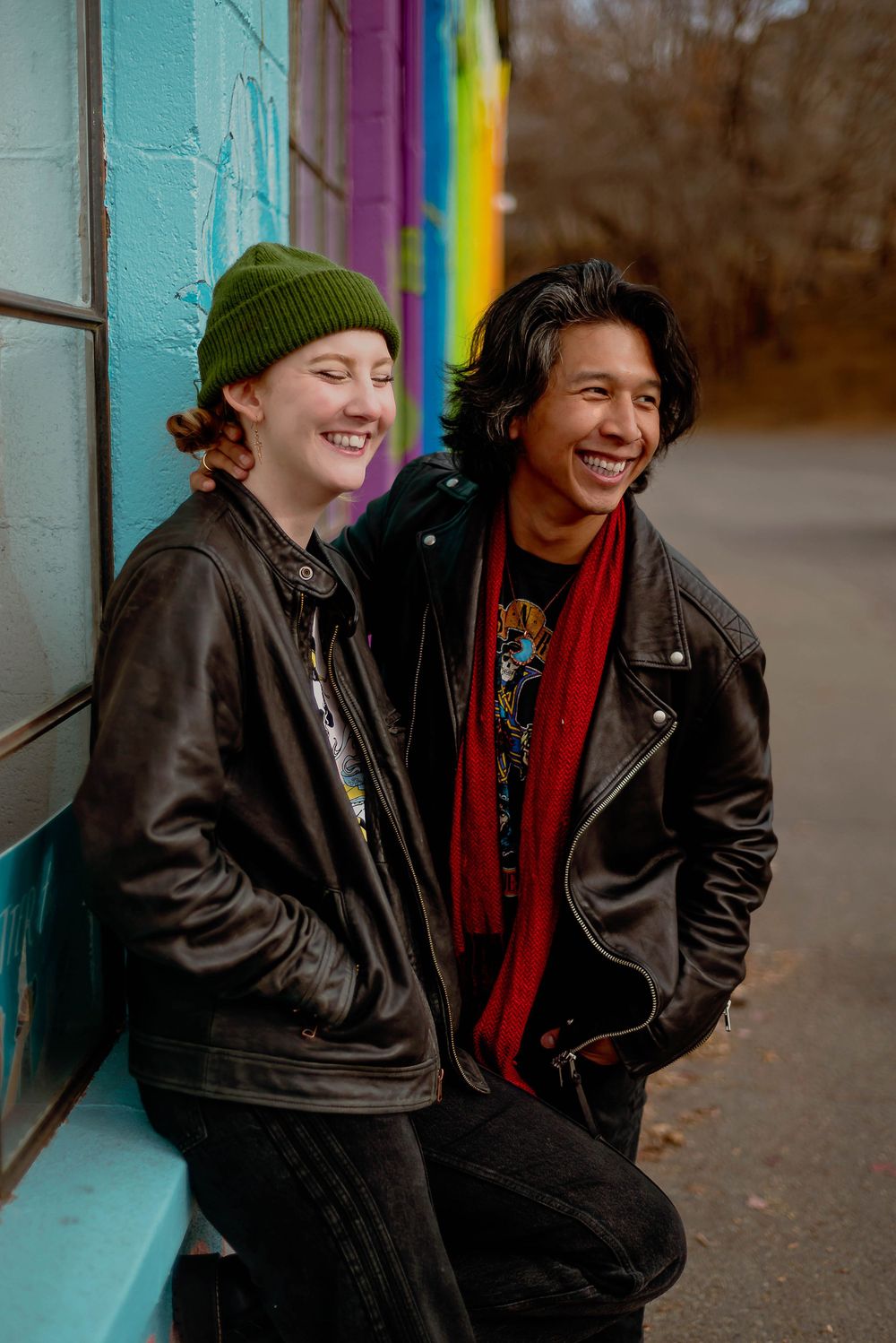 Two young friends laughing together while leaning against a turquoise wall, wearing winter clothes and leather jackets.
