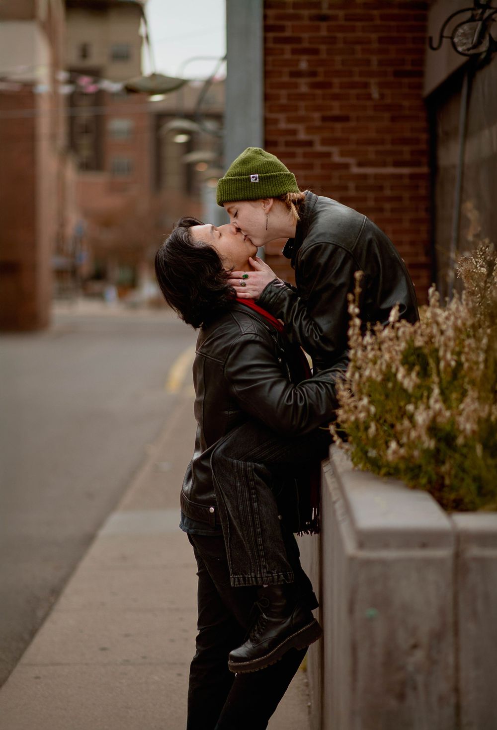 A tender romantic moment shared between two people in black winter clothing against a brick wall background.