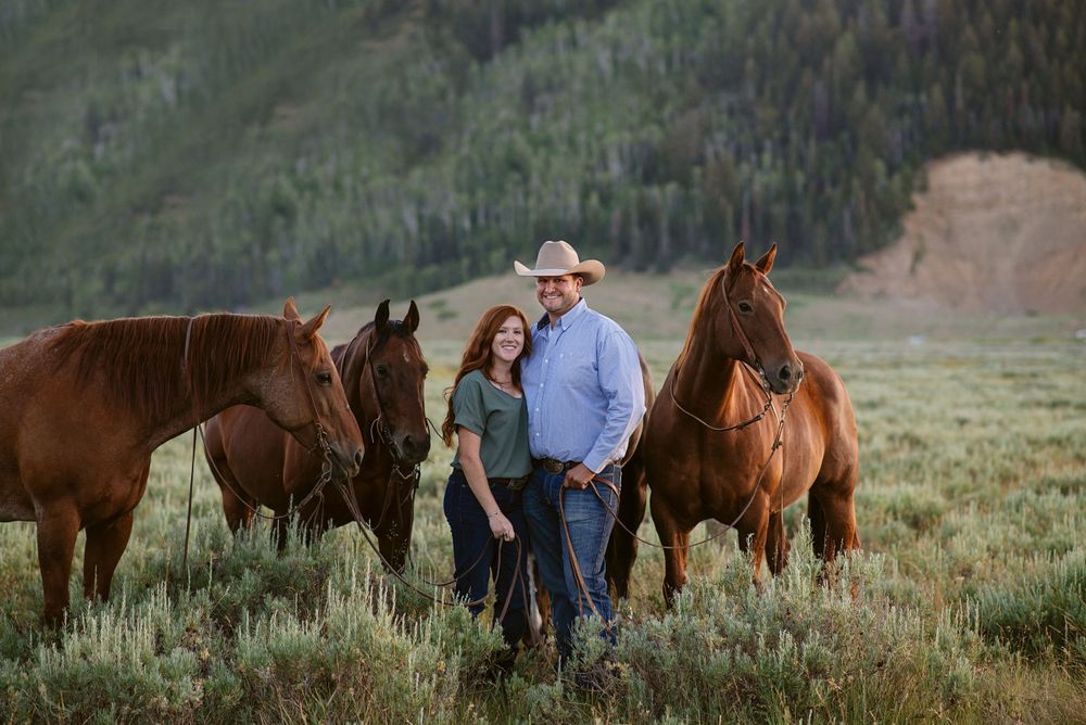 A couple stands with three brown horses in a grassy mountain meadow at dusk.