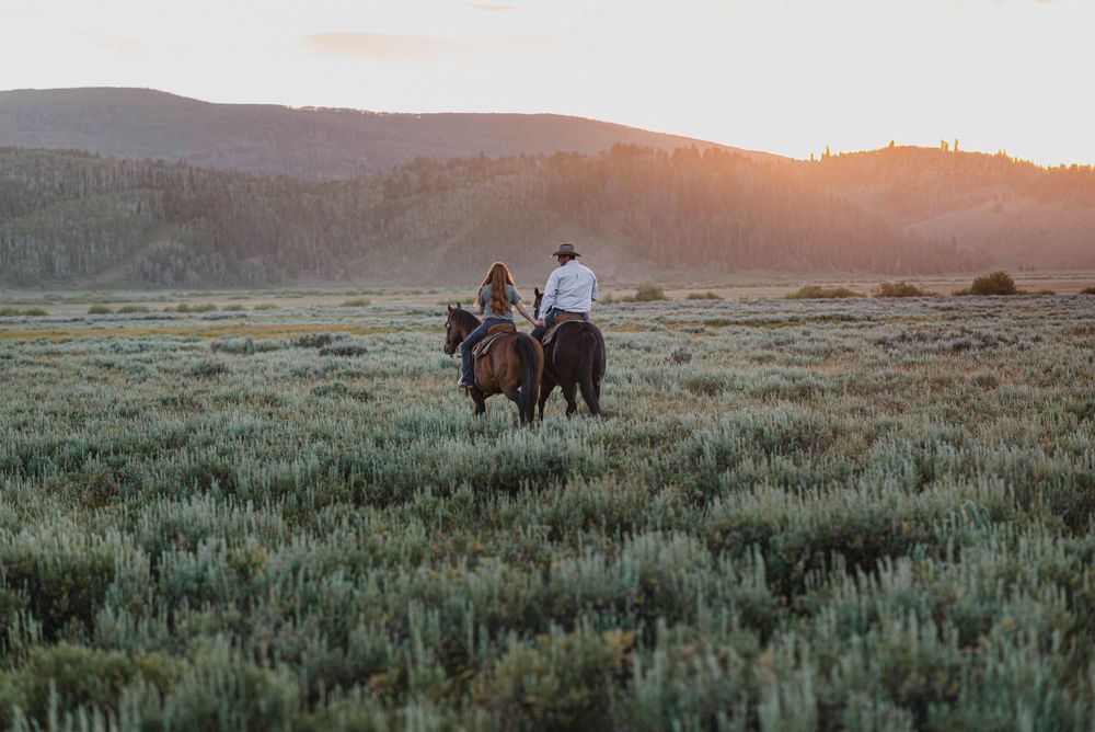 Two riders on horseback traversing a sage-covered field at sunset in the mountains.