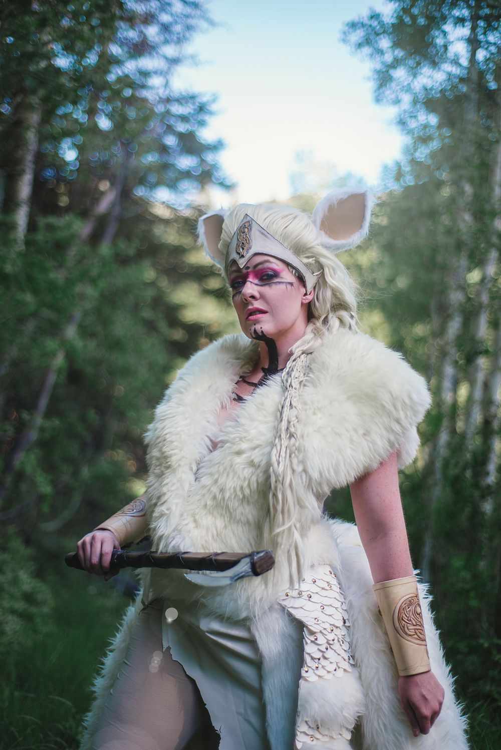 A cosplayer in a furry white costume with animal ears poses in a forest setting.