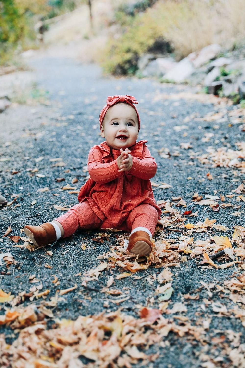 Small child in red outfit sits on ground playing with autumn leaves on a path.