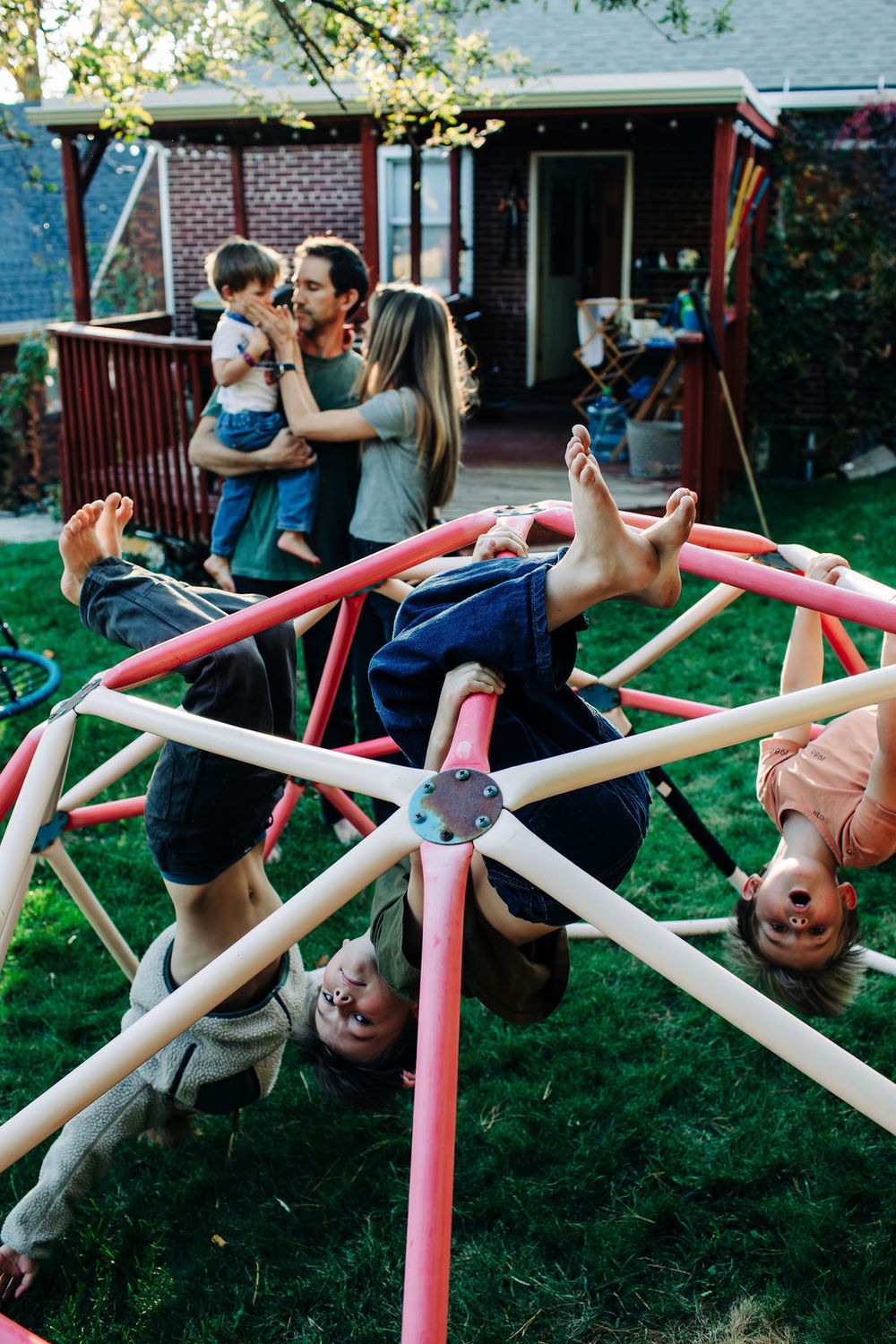 Children play on a colorful spinning playground equipment in a backyard while others gather on a deck in the background.
