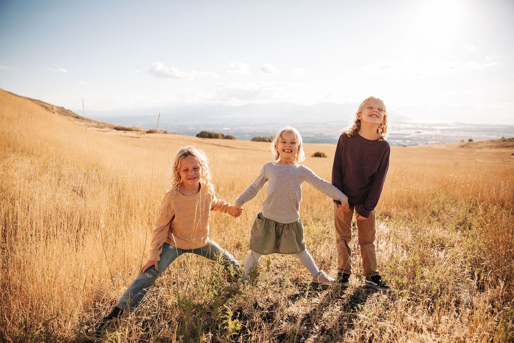 Three children hold hands and laugh together while playing in a golden field at sunset.