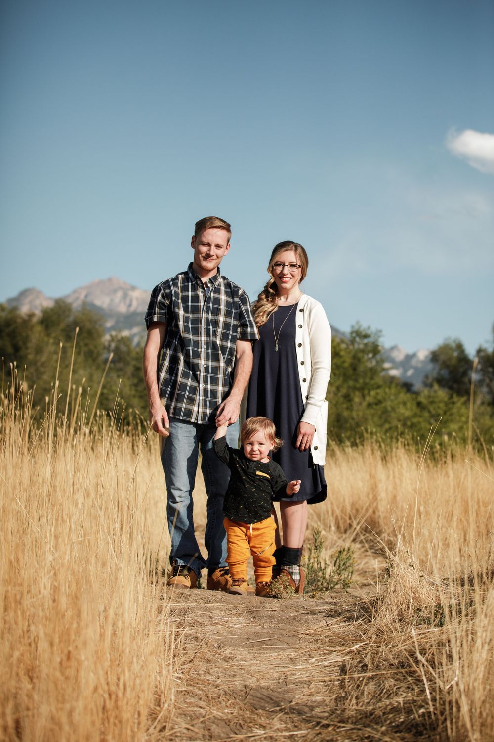 Family walking through tall golden grass in autumn field against mountain backdrop during sunset.