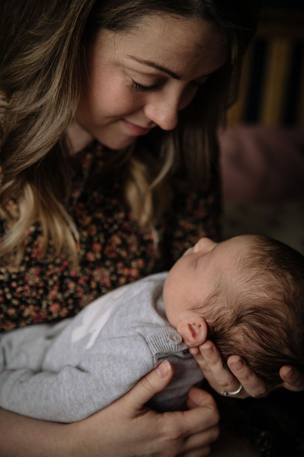 Mother in floral dress tenderly holding sleeping newborn in grey outfit.