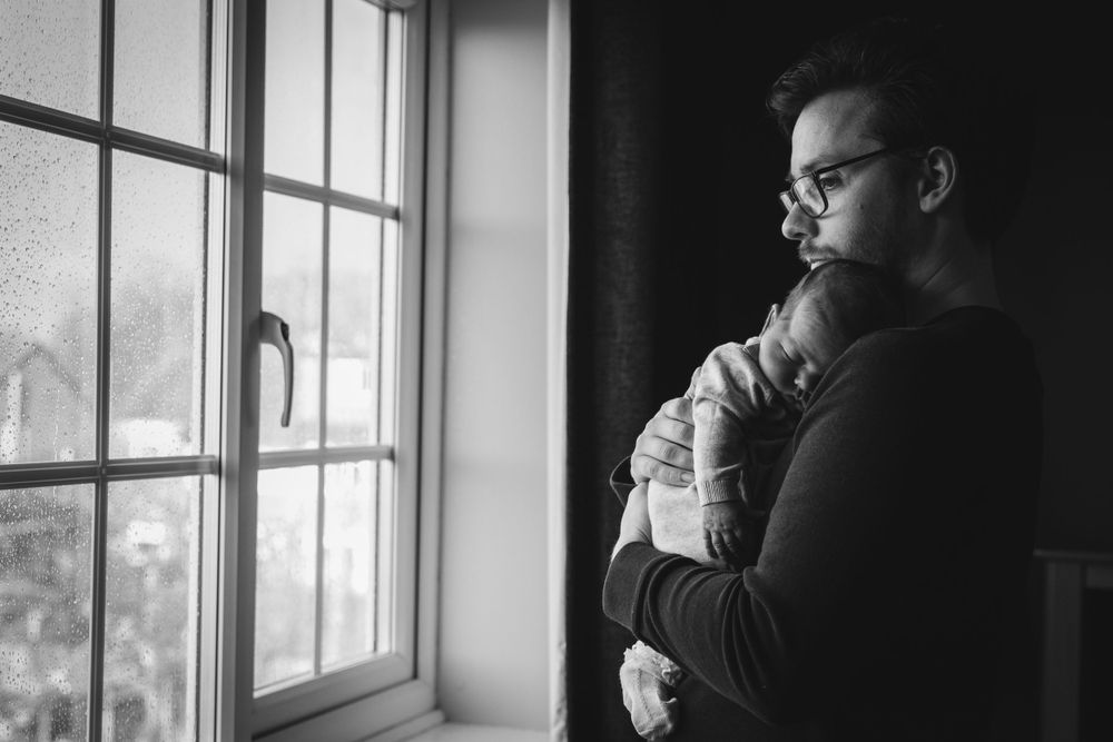 Parent holding sleeping infant while gazing out window in moody black and white.
