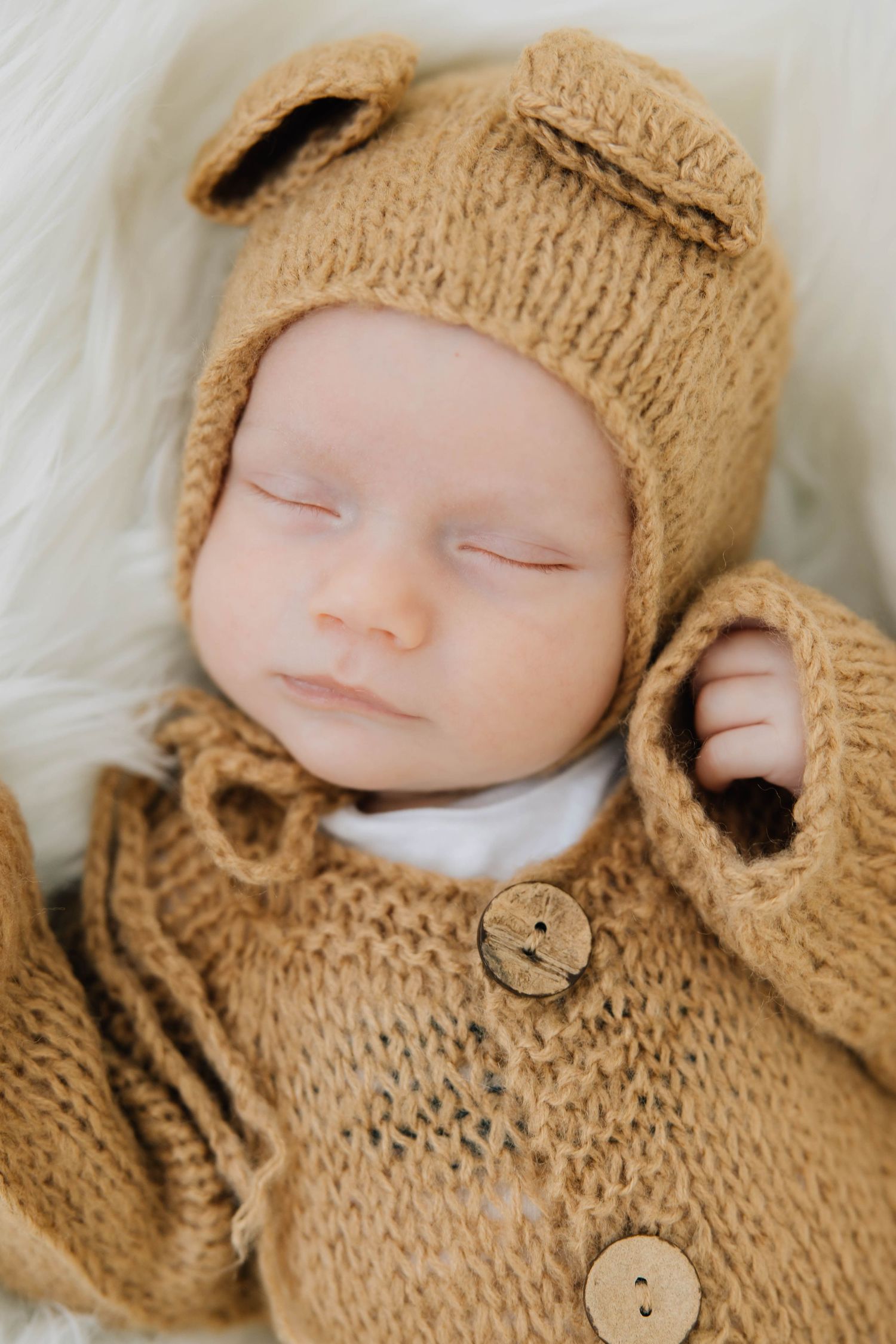 Sleeping newborn baby wearing a knitted tan bear bonnet and matching sweater outfit on a white fur blanket.