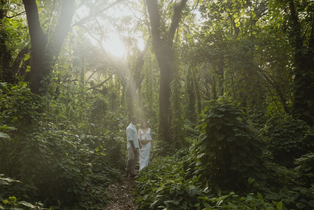 Couple in white attire walking through a lush green forest with sunlight streaming through tall trees.