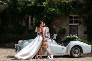 Wedding couple poses with vintage car outside historic stone building.