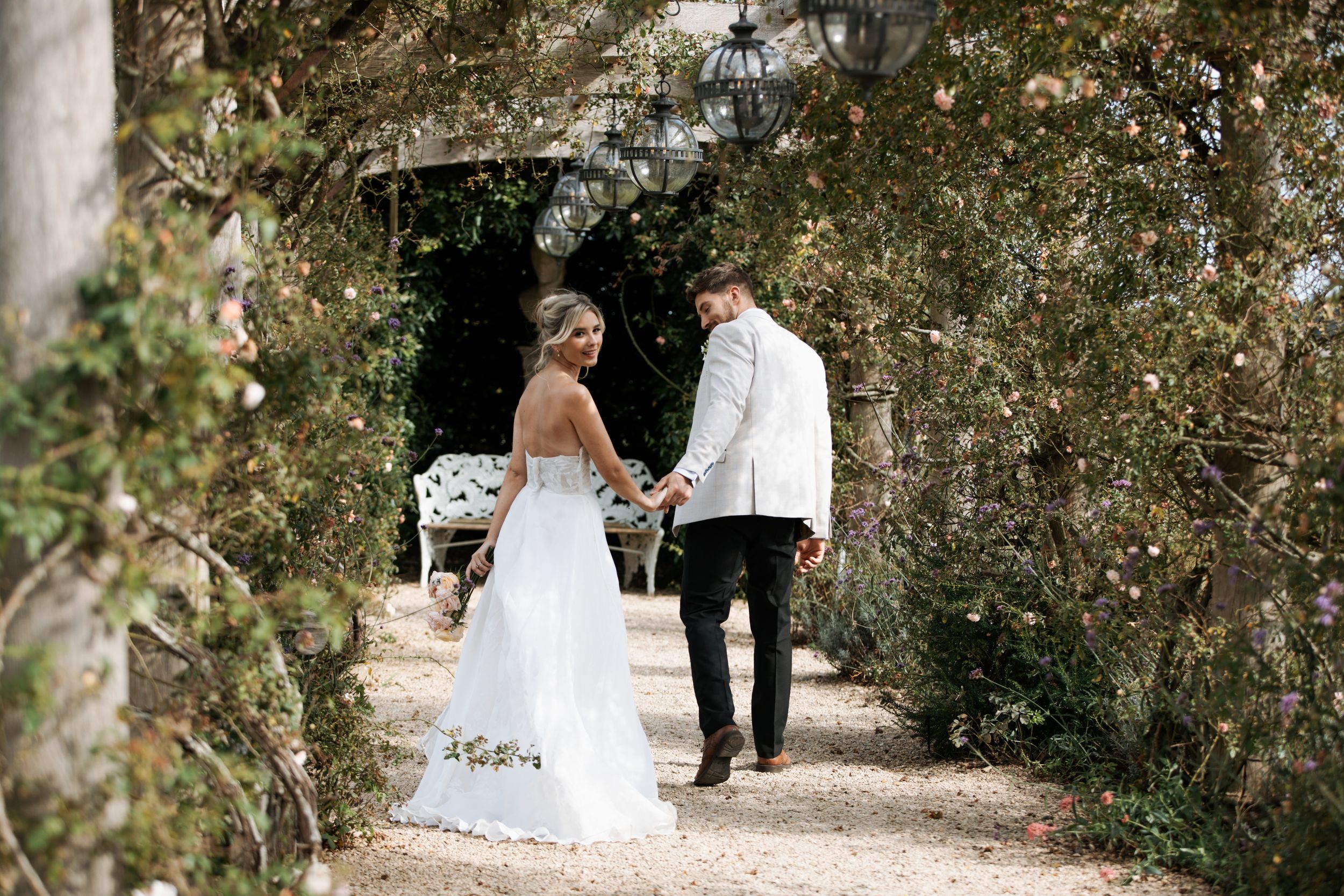 Newlyweds walk along a tree-lined path during their romantic outdoor wedding photoshoot.