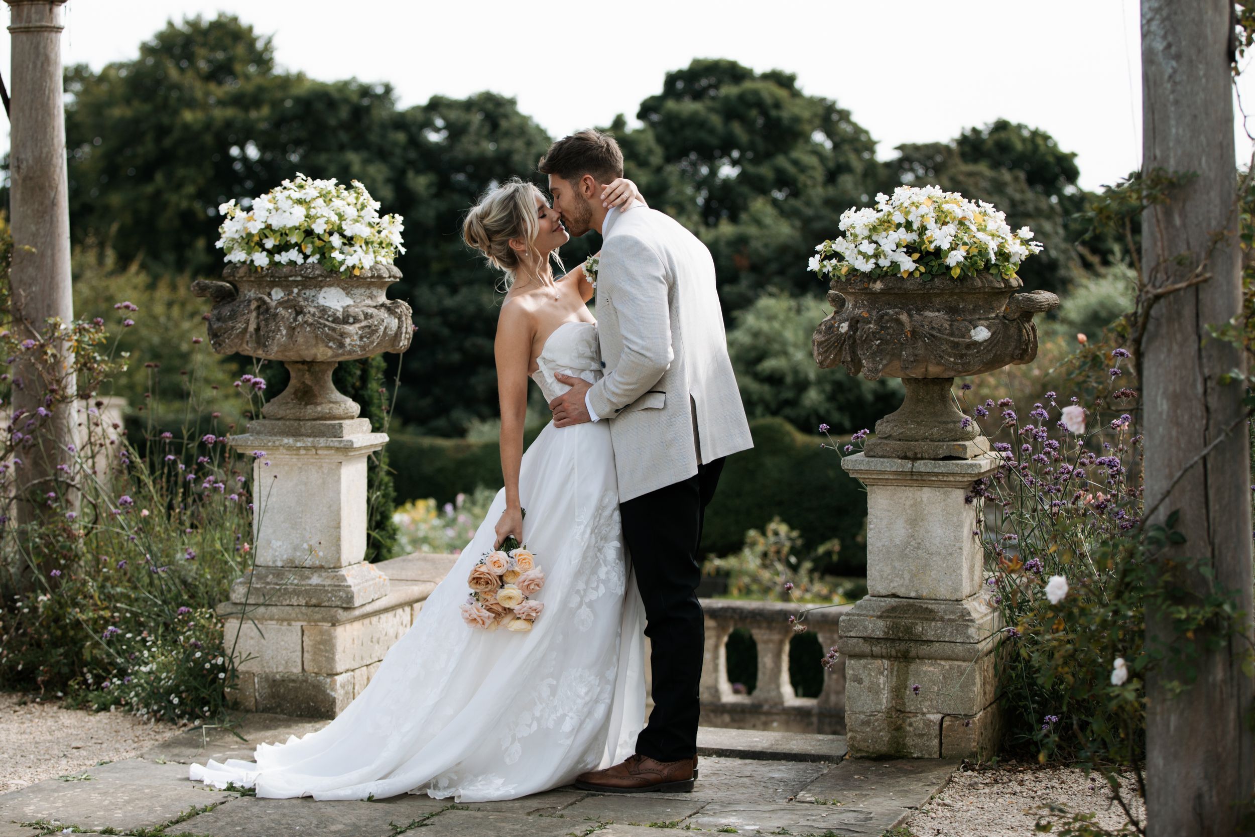Newlyweds pose romantically among classical stone pillars and urns in a formal garden setting.
