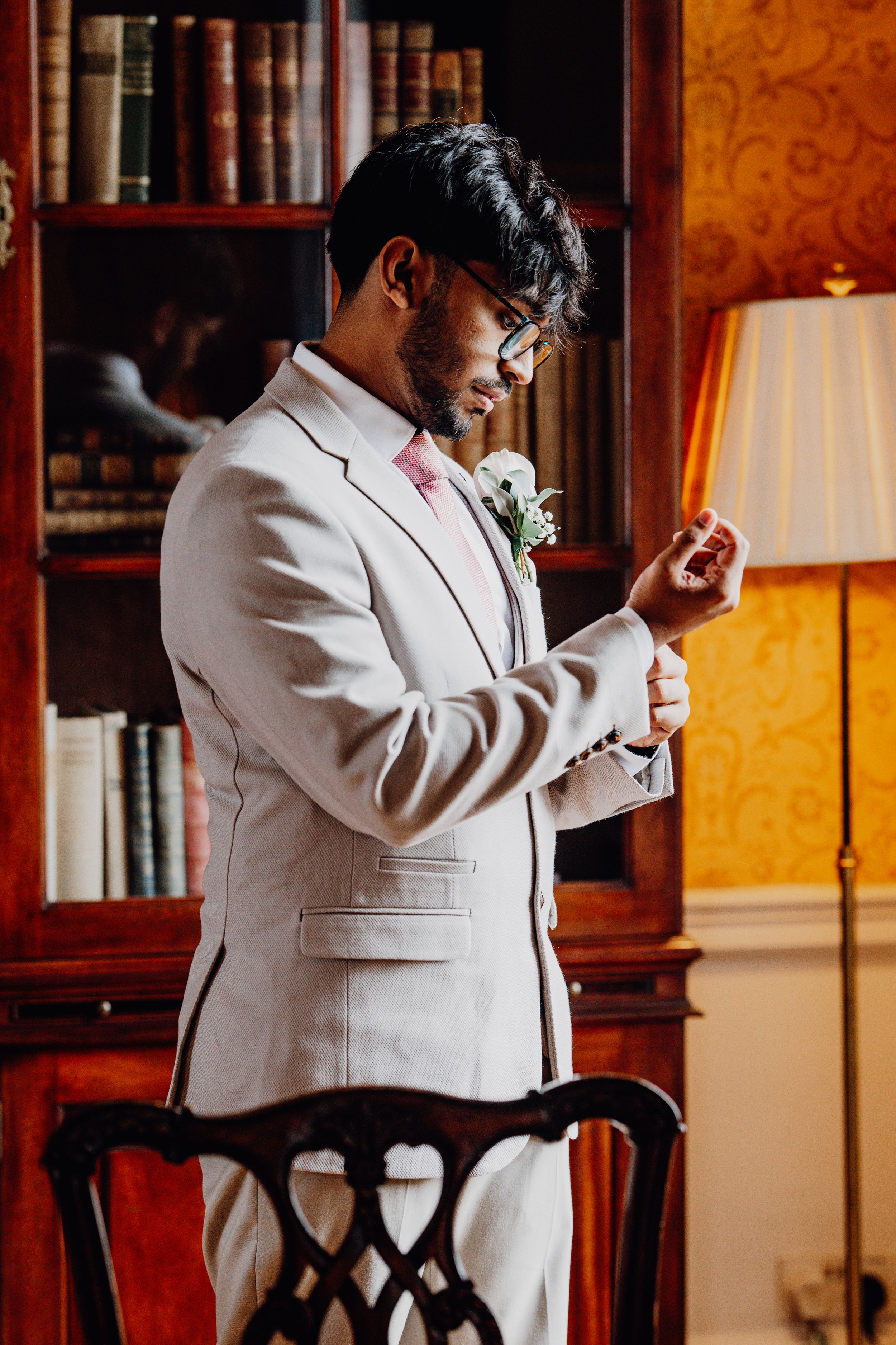 Elegant groom adjusts his tie and cufflinks in a warmly lit room with wood paneling.