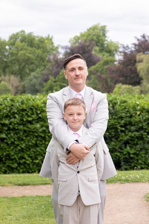 A playful sequence of a child and adult in light gray suits posing together outdoors against a green hedge background.