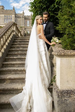 Couple poses on grand stone staircase surrounded by manicured gardens and historic architecture.