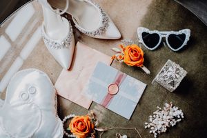 Wedding accessories including orange roses, jewelry, and invitation cards arranged on beige surface.