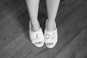 Feet wearing white bridal slippers on dark wooden flooring.
