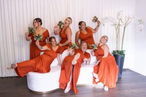 Wedding party wearing vibrant orange bridesmaid dresses pose together in a light-filled venue.