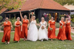 Wedding party in orange dresses and white gowns pose together on a grassy lawn.