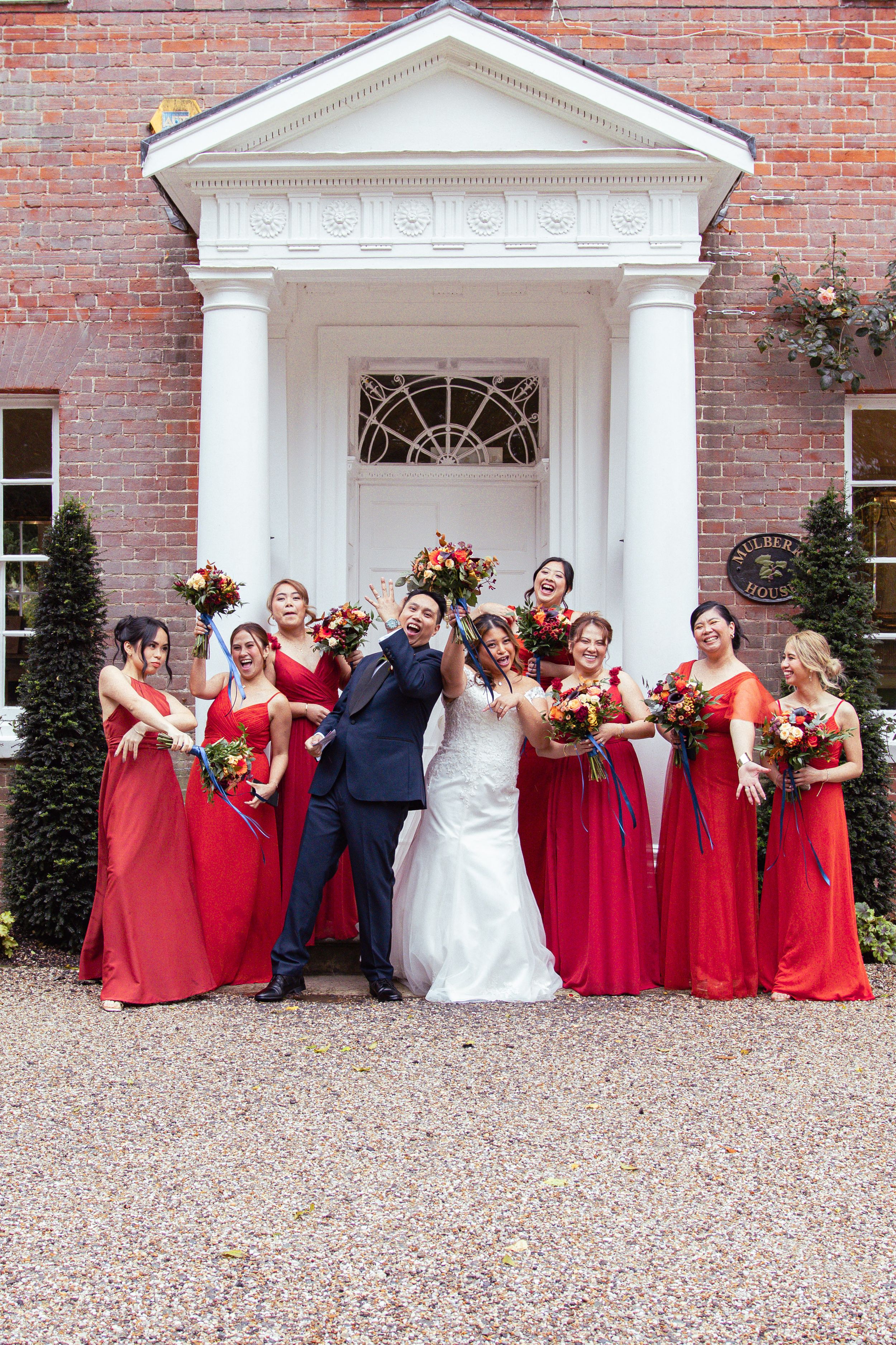 Bridal party in red dresses poses creatively along a brick wall with white columned entrance.