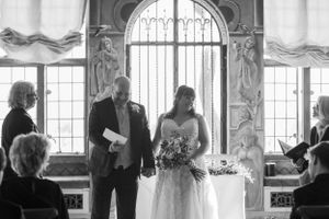 Wedding ceremony taking place in elegant room with large arched windows.