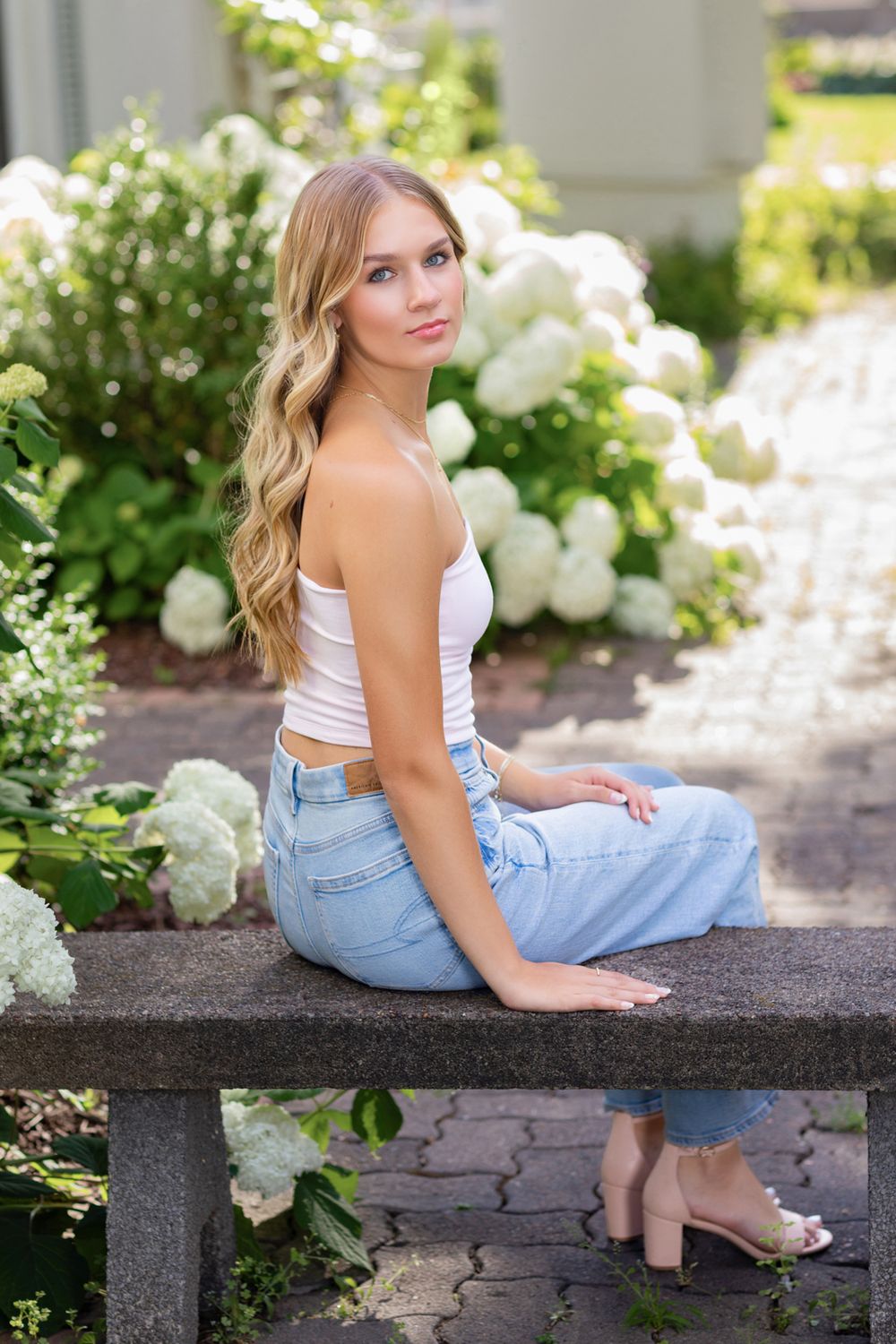 Person in casual summer outfit sitting on stone bench surrounded by white hydrangea bushes.