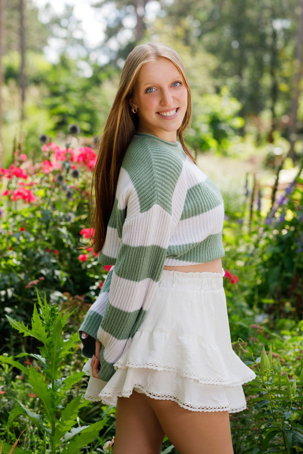 A person in a sage green and white striped sweater poses in a garden with blooming red flowers in the background.