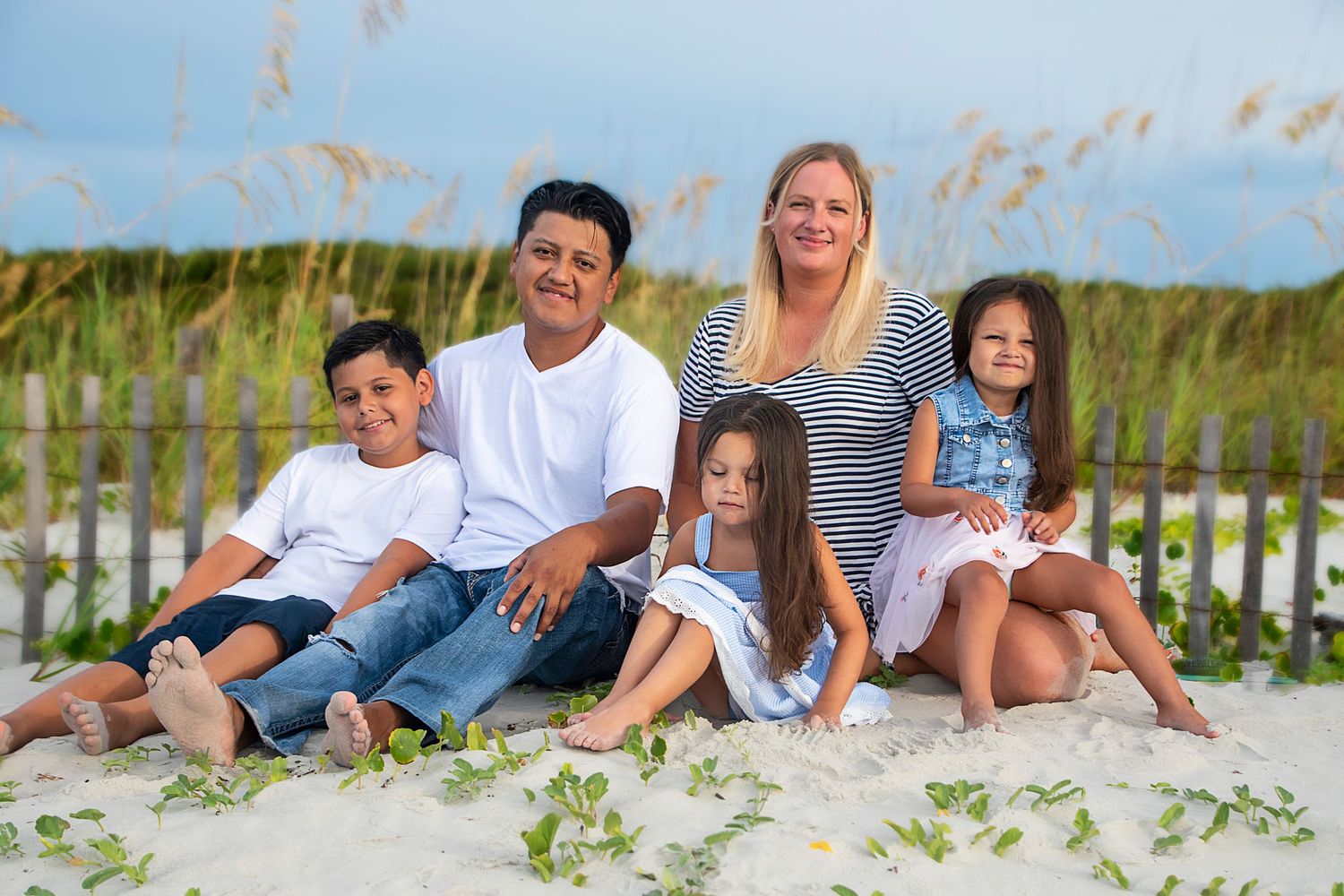 Beach family portrait with everyone sitting casually in the sand near sea oats.