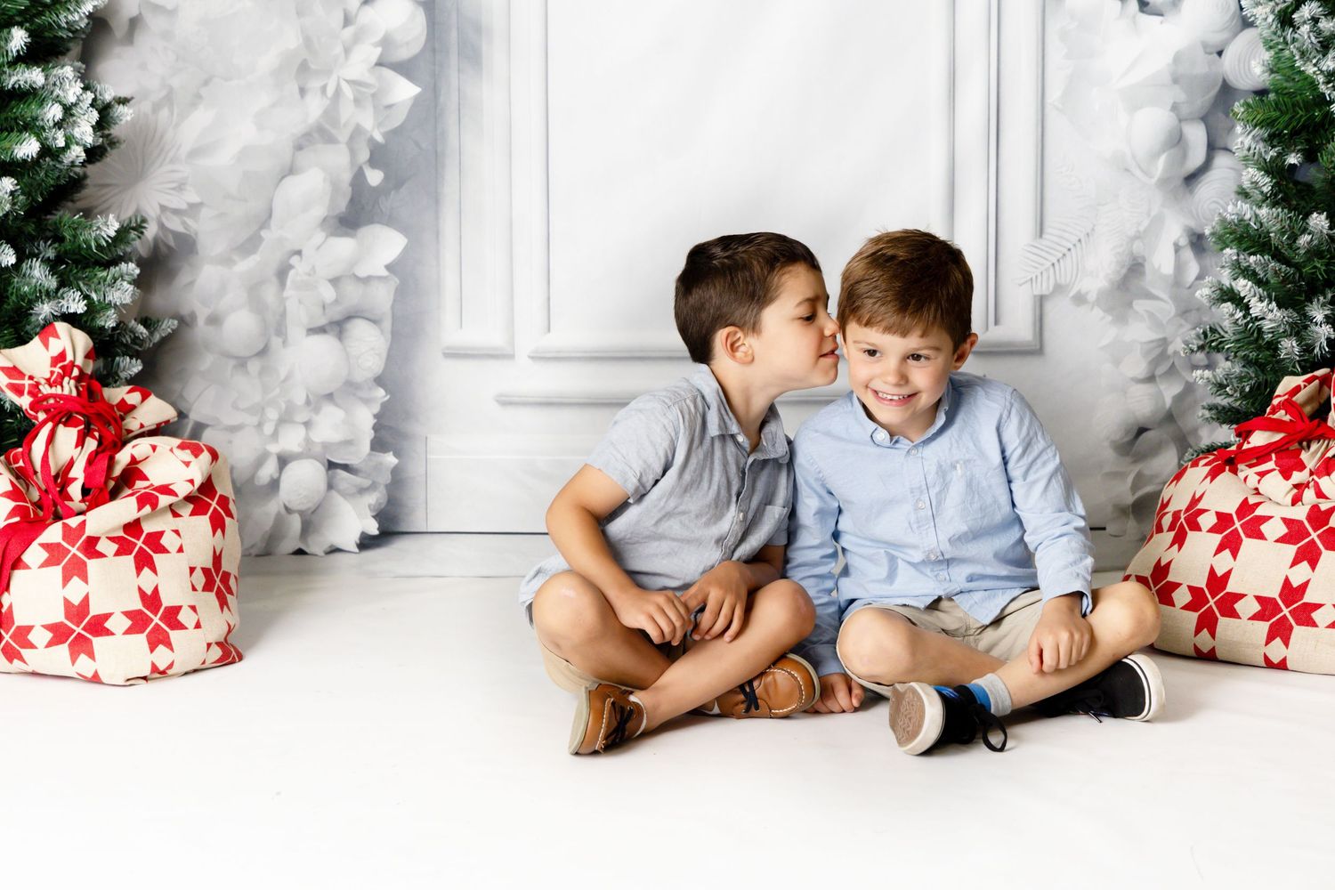 Two kids in light blue shirts share a laugh while seated against a white wall with red holiday gifts nearby.