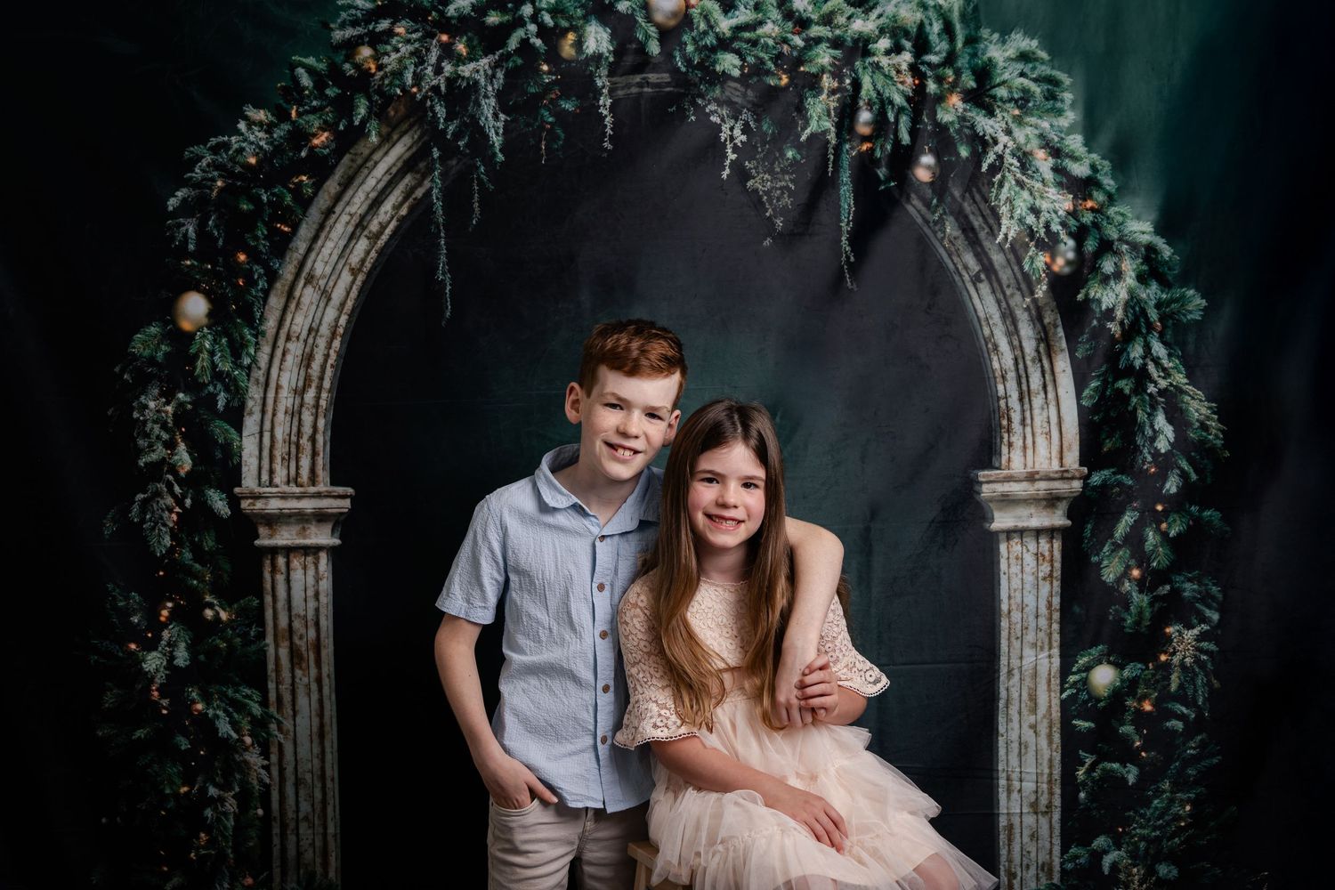 Two young people pose under an ornate stone archway decorated with lush greenery and floral arrangements.