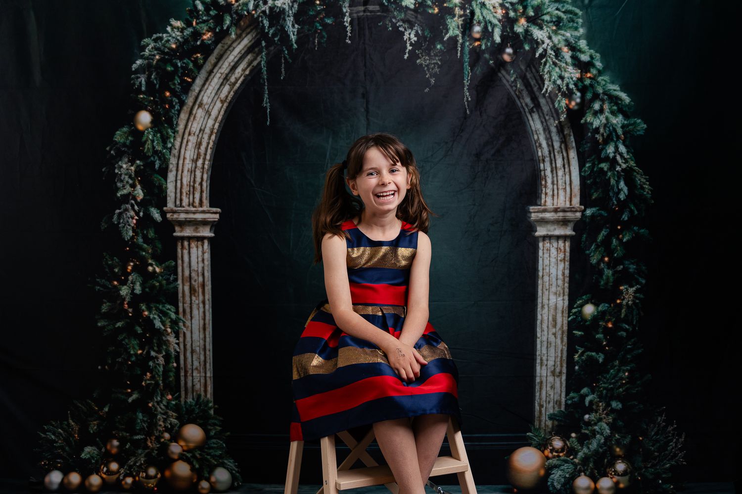A child in a red and black dress sits in front of a gothic archway decorated with vines and flowers in a dark studio.