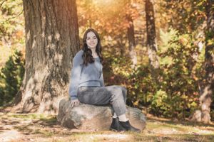 A woman in a gray sweater sitting on a rock surrounded by fall foliage.
