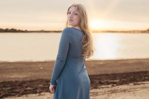 Woman in blue dress stands by water during sunset.