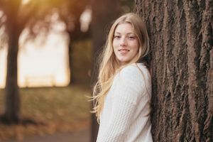 Woman in cream sweater leaning against tree trunk in autumn setting.