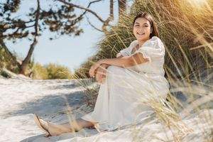 A person in a white dress sits on sandy beach dunes during golden hour sunset.