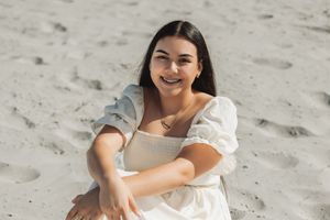 Person in white dress sitting casually on beach sand with a cheerful expression.