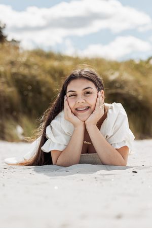 Person laying on beach sand with hands framing face in playful pose.