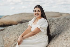 Person in white dress sitting on coastal rocks with ocean view background.