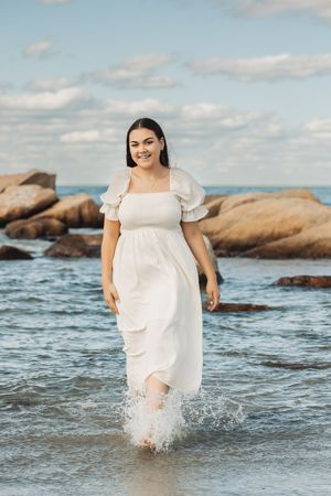 Person in white dress wading through ocean waves near rocky shore.